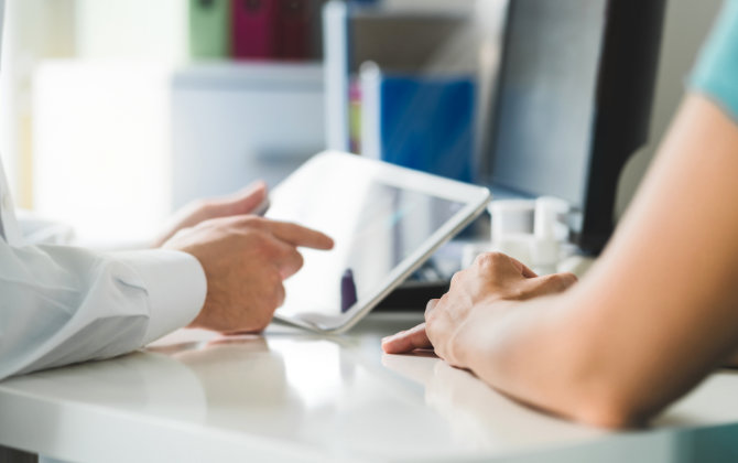 A doctor is looking at a tablet while talking to a patient.