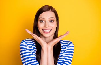 A woman with long brown hair and a striped shirt smiles broadly with her hands placed under her chin, set against a yellow background.