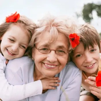 A smiling elderly woman with glasses is embraced by two children, each with a red flower in their hair.