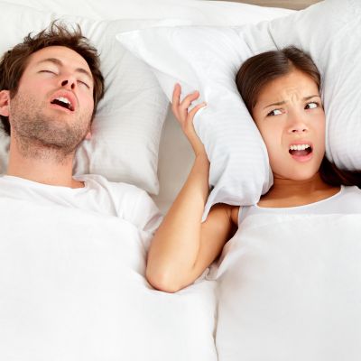 A man is sleeping and snoring while a woman covers her ears with a pillow, looking annoyed.