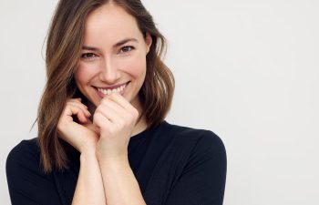 Woman with shoulder-length hair smiling and holding her hands near her face, wearing a black top against a plain background.