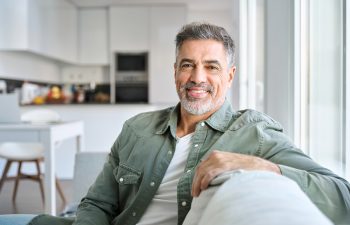 A middle-aged man with gray hair and a beard smiling while sitting on a couch in a bright modern living room.