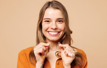 A young, happy woman with long brown hair smiles broadly at the camera while holding a set of clear dental aligners in front of her. She is wearing an orange button-up shirt and is set against a plain, light brown background.