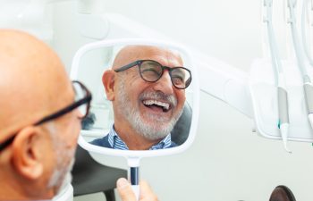 A smiling older man with glasses and a gray beard looks at his reflection in a hand mirror, checking his teeth in a dentist's office.