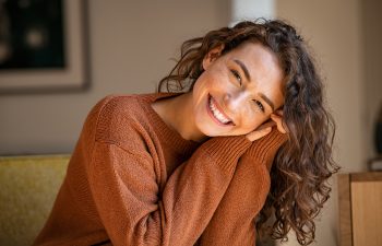 A woman with curly brown hair wearing a rust-colored sweater smiles and rests her head on her hands while seated indoors.