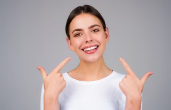 A smiling young woman with brown hair pulled back in a white shirt points to her white teeth with both index fingers, set against a gray background.