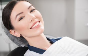 A young woman smiles brightly while reclining in a dental chair, wearing a white paper bib.