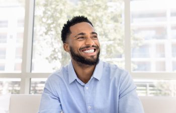 A happy man with a short beard and dark hair, wearing a light blue button-down shirt, smiles while looking off to the side. He is sitting indoors in front of a large window with bright natural light