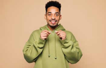 A smiling young man in a green hoodie holding a clear orthodontic aligner with both hands against a beige background.